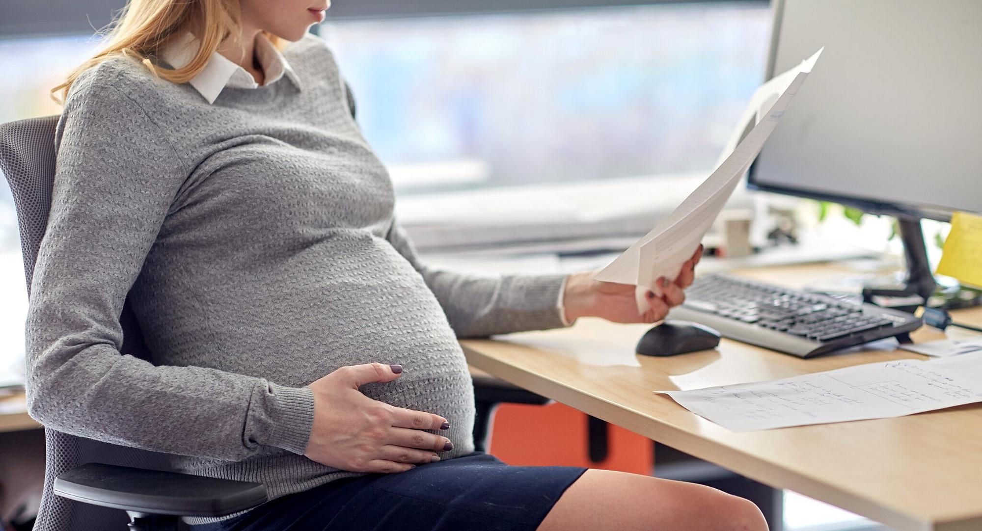 Eine schwangere Frau sitzt an ihrem Arbeitsplatz und hält Zettel in der Hand.