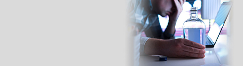 Ein Mann sitzt mit Alkoholflasche vor dem Laptop und sieht bedrückt aus.