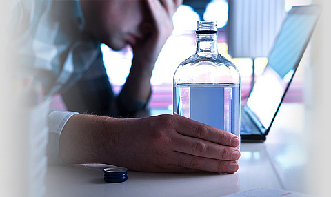 Ein Mann sitzt mit Alkoholflasche vor dem Laptop und sieht bedrückt aus.