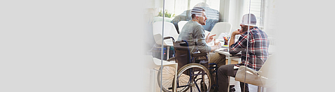 Ein Mann im Rollstuhl sitzt mit einem anderen Mann am Tisch und bespricht sich.