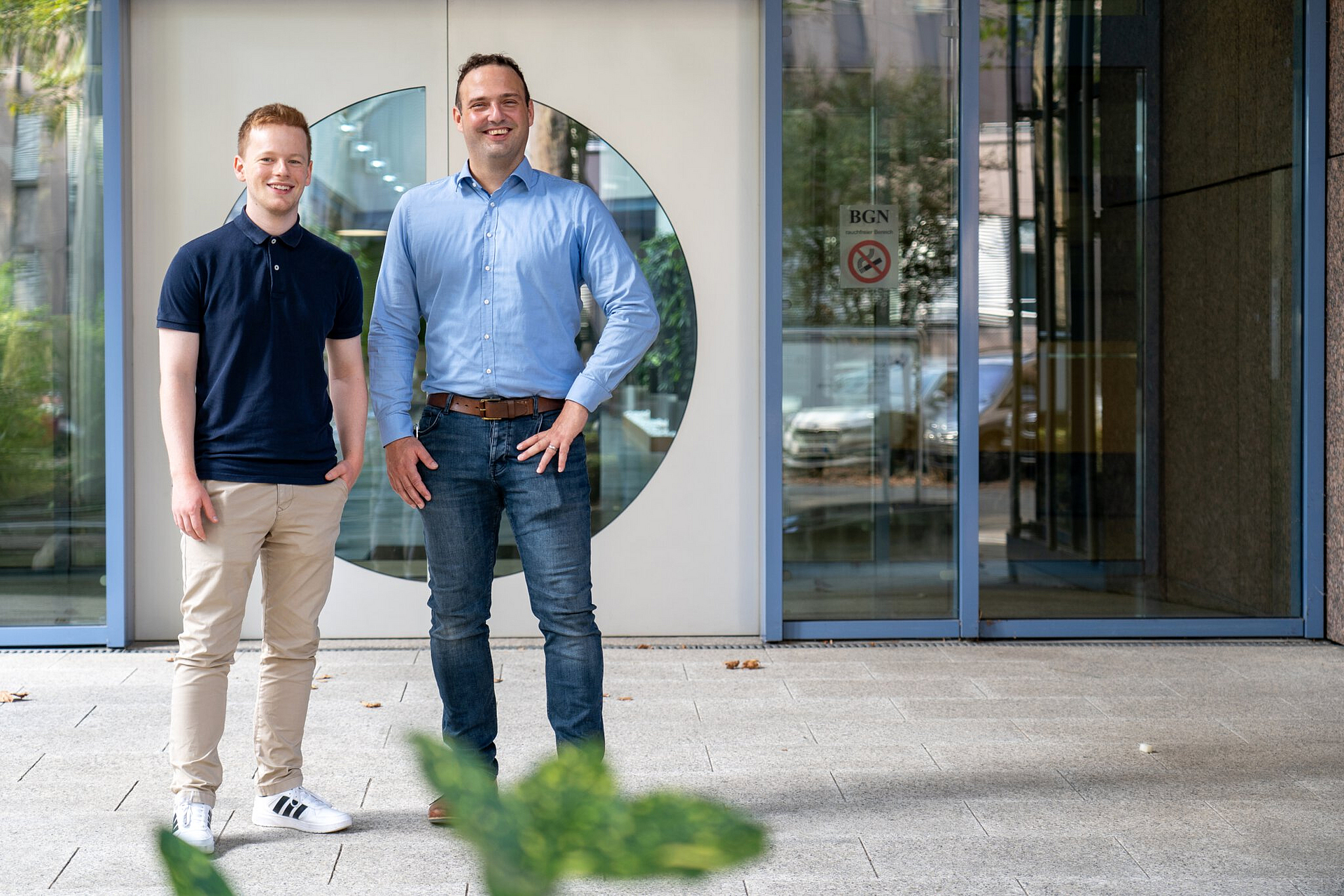 Von links: Jonas Brenneis und Dr. Andre Becker stehen lächelnd vor der Eingangstür des BGN-Schulungszentrums