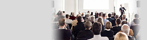 Blick in einen Raum mit mehreren Personen von hinten die auf einen Referenten im Hintergrund blicken.
