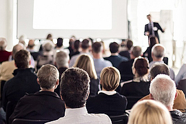 Blick in einen Raum mit mehreren Personen von hinten die auf einen Referenten im Hintergrund blicken.