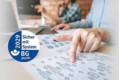 Ein Mann sitzt am Schreibtisch und zeigt auf ein Blatt Papier mit einem Diagramm, daneben das AMS-Gütesiegel.