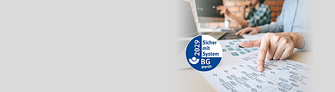 Ein Mann sitzt am Schreibtisch und zeigt auf ein Blatt Papier mit einem Diagramm, daneben das AMS-Gütesiegel.