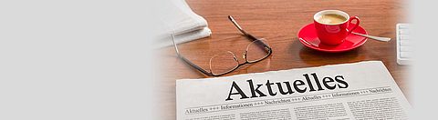 Auf einem Tisch liegt eine Zeitung mit dem Title Aktuelles, eine Brille und ein Kaffeebecher.