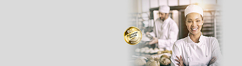 Bäckerin lächelt in die Kamera, im Hintergrund arbeitet ein Bäcker mit Teig.