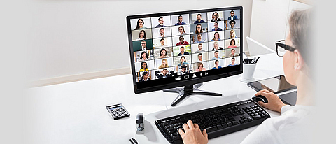 Frau sitzt am Schreibtisch, der PC-Bildschirm zeigt eine Videokonferenz.