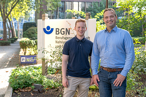 Von links: Jonas Brenneis und Dr. Andre Becker stehen lächelnd vor einem Schild der Berufsgenossenschaft Nahrungsmittel und Gastgewerbe (BGN)
