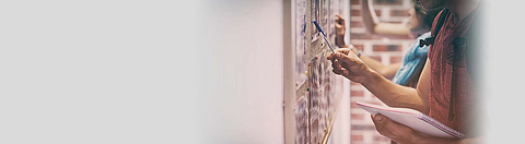 Zwei Personen stehen vor einer Pinnwand und zeigen auf einen Aushang.