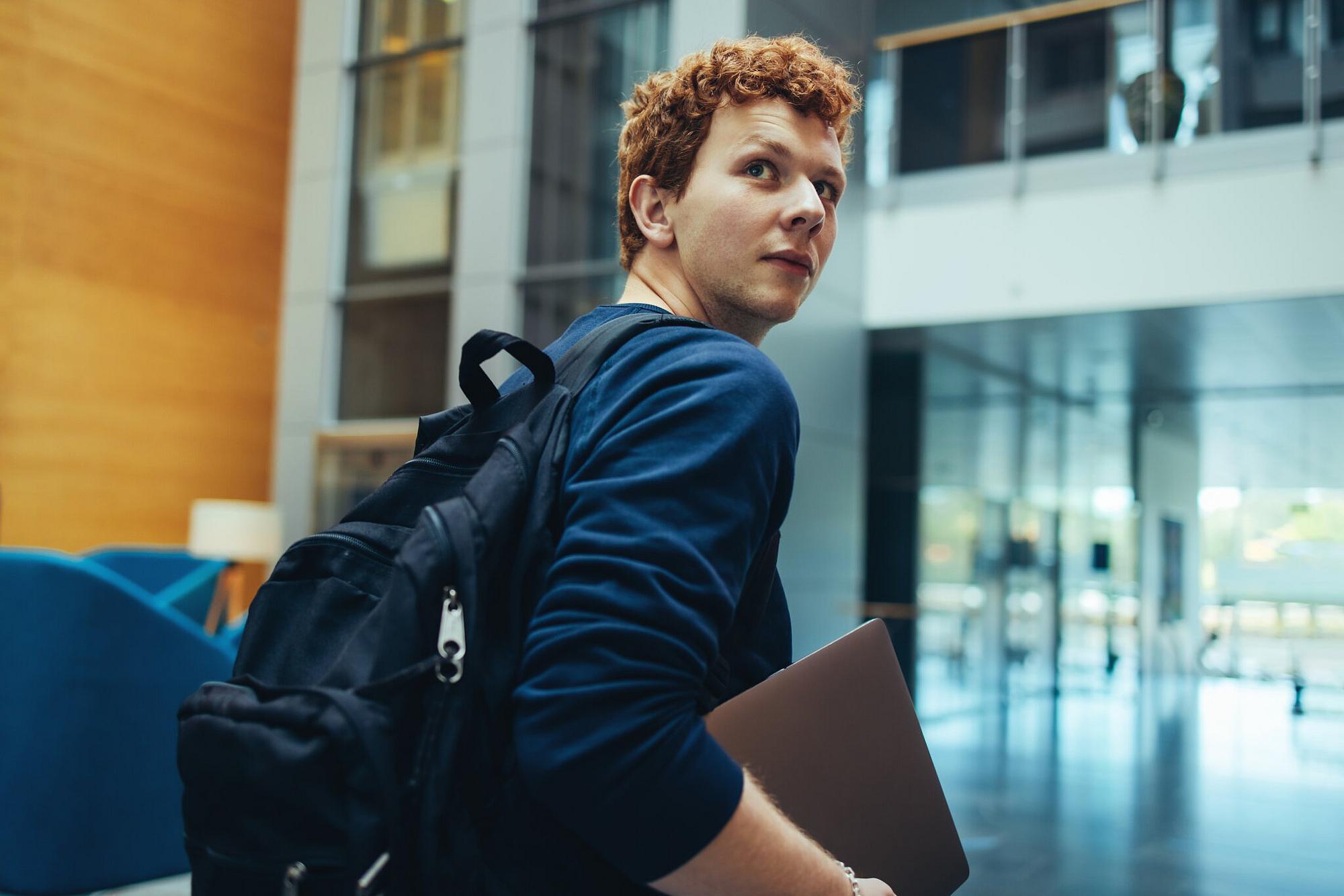 Ein junger Mann mit roten Haaren, dunkelblauem Pullover und Rucksack trägt einen Laptop unter dem Arm durch ein großes Gebäude mit Glasfronten und Aufzügen.