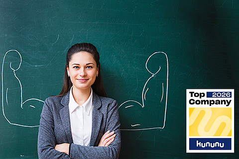 Eine junge Frau steht vor einer Tafel, auf der Tafel sind Arme mit großen Muskeln aufgemalt.