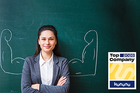 Eine junge Frau steht vor einer Tafel, auf der Tafel sind Arme mit großen Muskeln aufgemalt.
