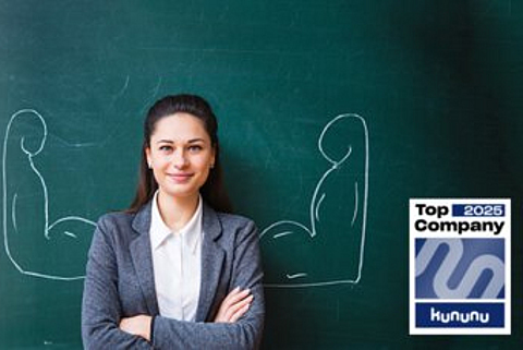 Eine junge Frau steht vor einer Tafel, auf der Tafel sind Arme mit großen Muskeln aufgemalt.