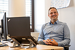 Dr. Andre Becker sitzt lächelnd an einem Schreibtisch vor einem Laptop und einem Monitor
