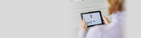 Frau schaut auf ein Tablet, das einen Arzt per Videochat zeigt.