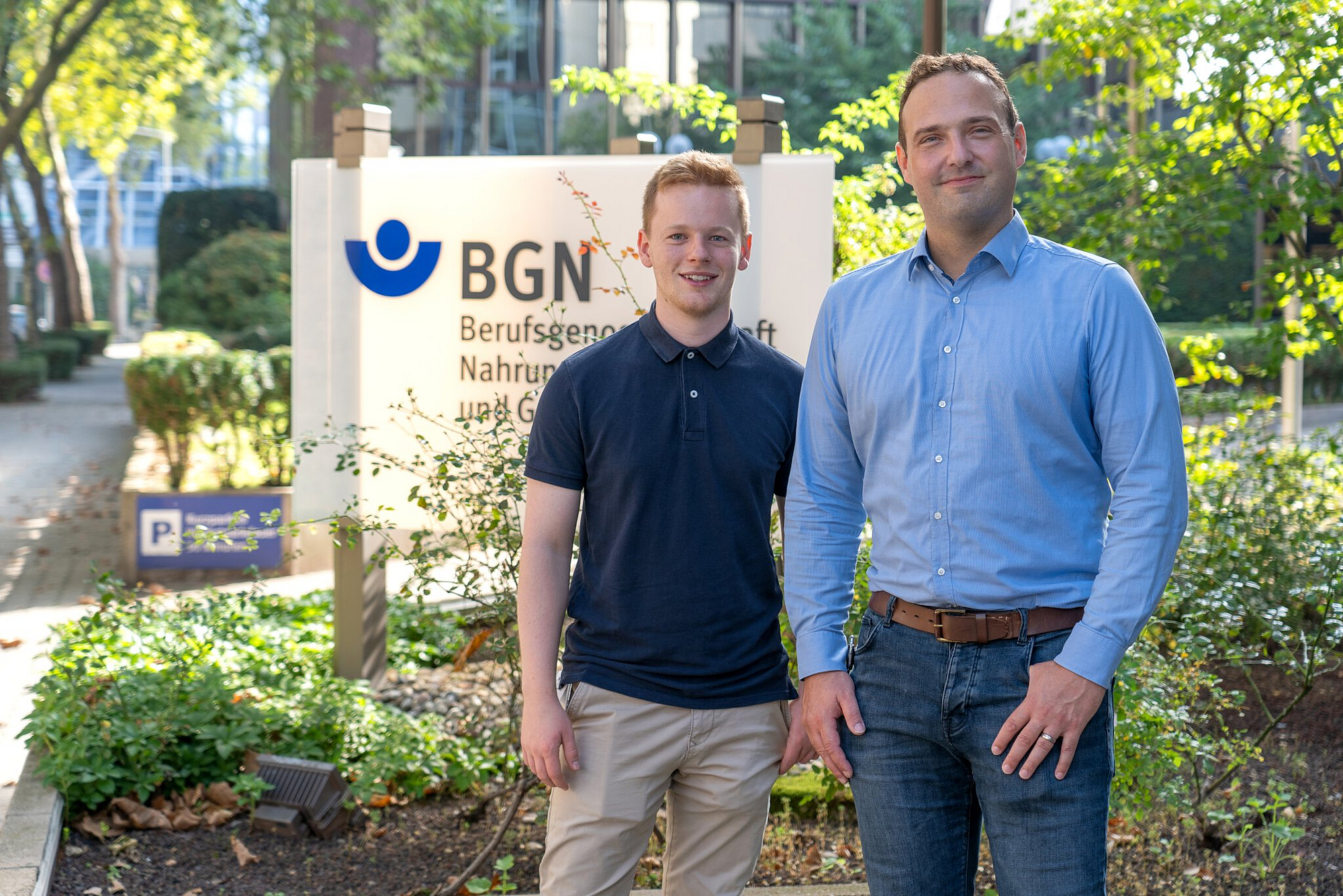 Von links: Jonas Brenneis und Dr. Andre Becker stehen lächelnd vor einem Schild der Berufsgenossenschaft Nahrungsmittel und Gastgewerbe (BGN)