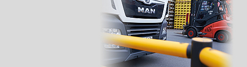 Ein LKW und ein Gabelstapler auf einem Betriebsgelände, im Vordergrund ein Geländer des Fußgängerwegs