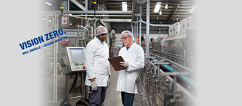 Zwei Männer stehen in einer Fabrikhalle und schauen auf ein Klemmbrett.