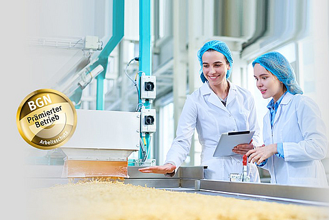 Zwei Mitarbeiterinnen in Schutzkleidung stehen an einem Laufband mit Nudeln und betrachten die Produkte.