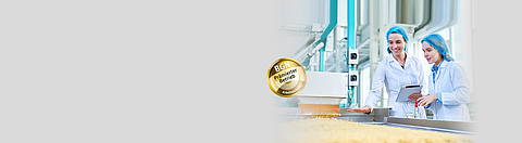 Zwei Mitarbeiterinnen in Schutzkleidung stehen an einem Laufband mit Nudeln und betrachten die Produkte.