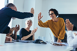 Mitarbeitende sitzen am Tisch und applaudieren, ein Mitarbeiter klatscht eine Mitarbeitende ab.