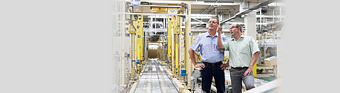 Zwei Männer stehen in einer Fabrikhalle, ein Mann zeigt auf etwas.