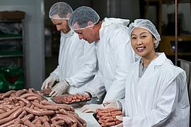 Zwei Mitarbeiter und eine Mitarbeiterin eines Fleischereibetriebes tragen Schutzkleidung und packen Wurstwaren ab