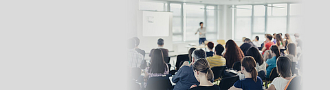 Publikum von hinten, im Hintergrund steht ein Mann und präsentiert etwas.