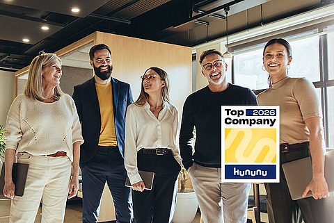 Fünf Kollegen stehen beieinander in einem Büro und lachen. Im Vordergrund das Kununu Top Company Logo.
