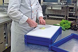 Ein Koch steht vor einem Schneidbrett und schneidet Gemüse, das Schneidbrett ist auf seine Höhe angepasst.