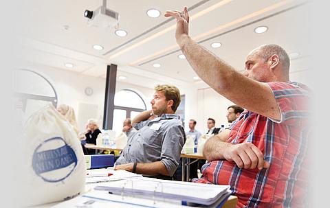 Mehrere Personen sitzen in einem Raum bei einem Seminar, ein Mann im Vordergrund hebt seine Hand, um sich zu melden.