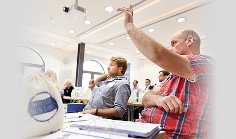 Mehrere Personen sitzen in einem Raum bei einem Seminar, ein Mann im Vordergrund hebt seine Hand, um sich zu melden.