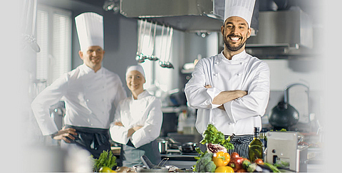 Ein Koch lacht in die Kamera, im Hintergrund stehen zwei Kollegen.