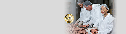 Eine Mitarbeiterin und zwei Mitarbeiter eines Fleischereibetriebs tragen Schutzkleidung und packen Wurstwaren ab.