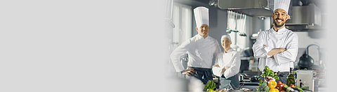 Ein Koch lacht in die Kamera, im Hintergrund stehen zwei Kollegen.