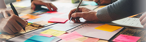 Verschiedene Menschen stehen um einen Tisch voller Dokumente und bunter Notizzettel und schreiben Ideen auf.