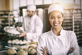 Eine Bäckerin lächelt in die Kamera während ihr Kollege im Hintergrund Teig vorbereitet