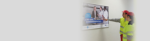 Zwei Mitarbeitende schauen und zeigen auf ein Plakat der Kampagne "kommmitmensch".