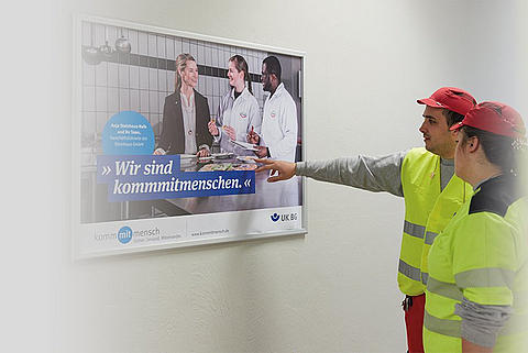 Zwei Mitarbeitende schauen und zeigen auf ein Plakat der Kampagne "kommmitmensch".