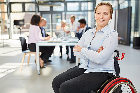 Junge Frau im Rollstuhl sitzend. Im Hintergrund findet eine Besprechung statt.