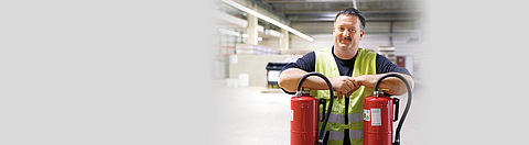 Ein Mann lehnt sich mit den Armen auf zwei Feuerlöscher und lächelt in die Kamera.