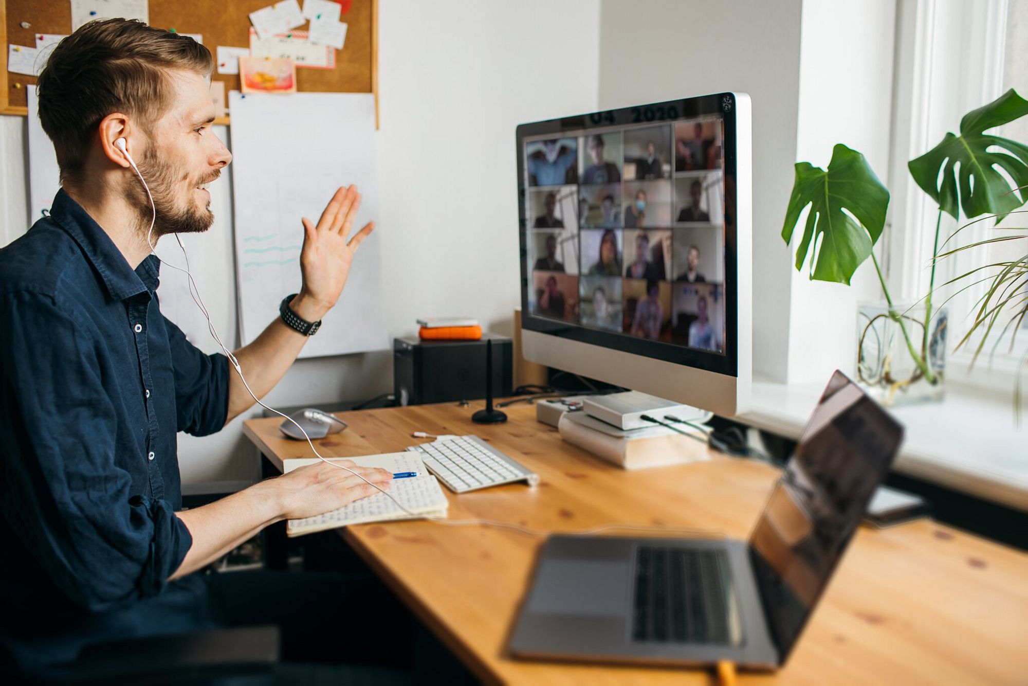 Ein Mann sitzt am Schreibtisch vor einem PC, auf dem Bildschirm läuft ein Videochat. Der Mann winkt in die Kamera.